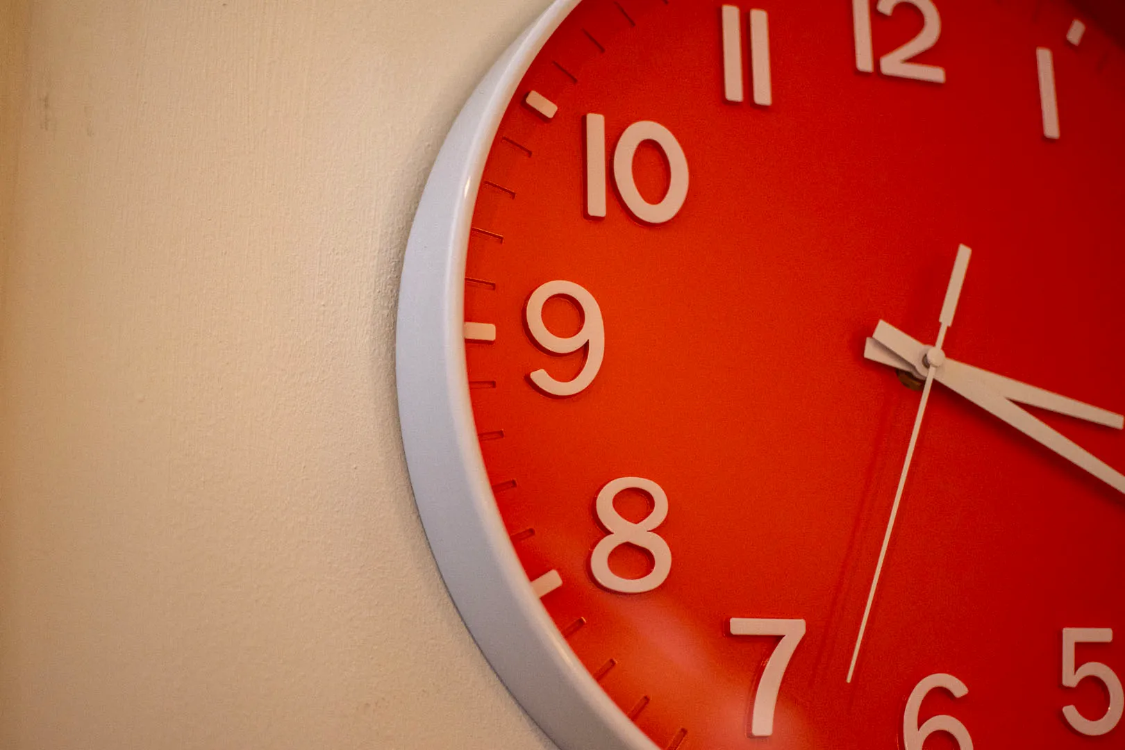 A close-up of a red and white analog clock with white numbers and hands, set against a beige wall.