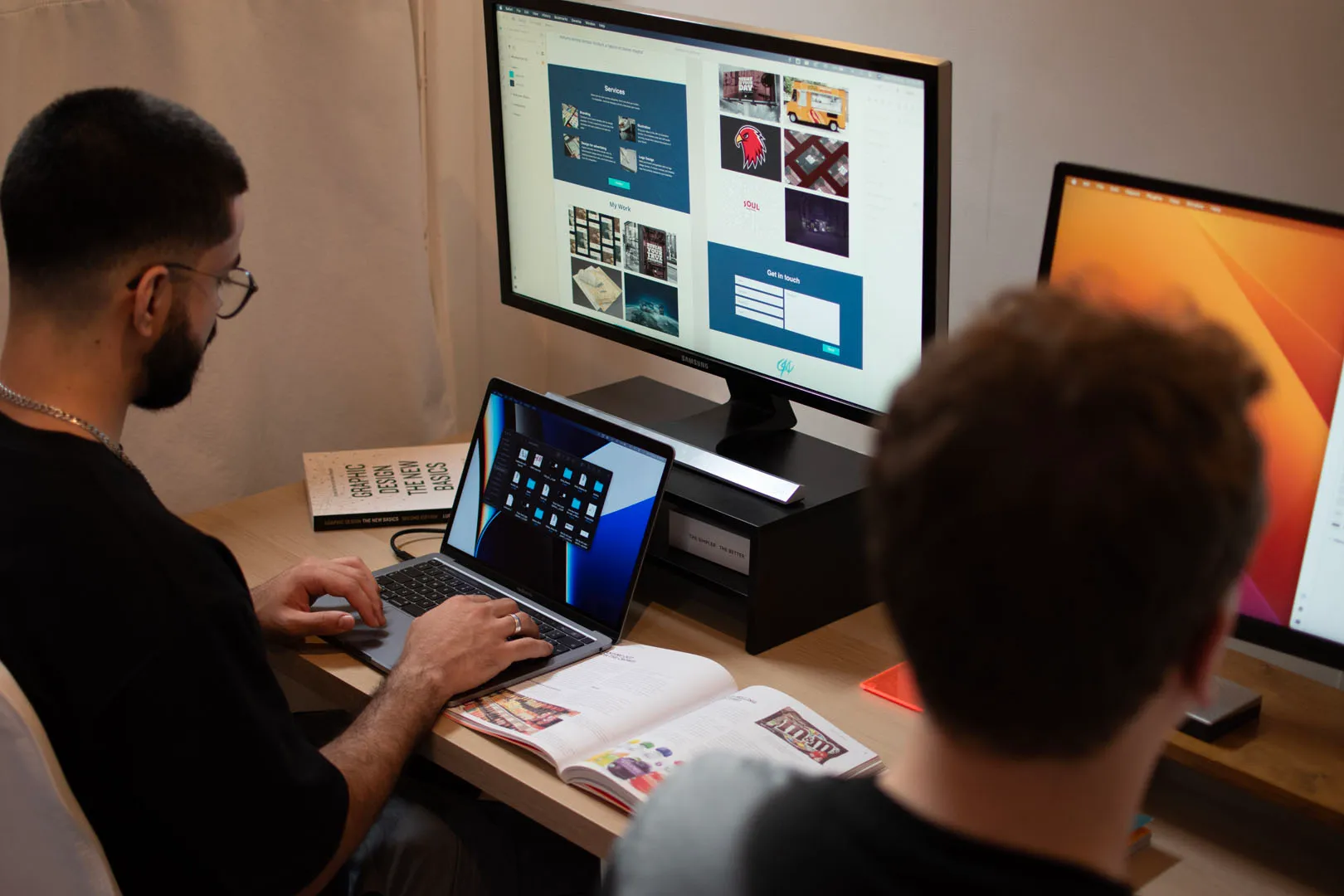 Two men sit at a desk with two computer monitors and a laptop, surrounded by books and a notebook. The man on the left works on a laptop, while the man on the right looks at the screens. The screens display a website design project, with a