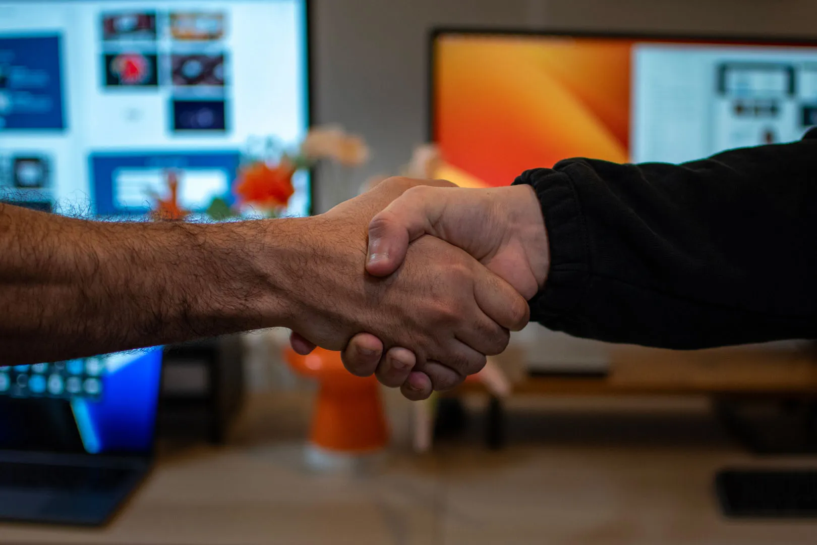 Two people shake hands across a desk in the Berlew studio, with design work visible on the monitors behind them.