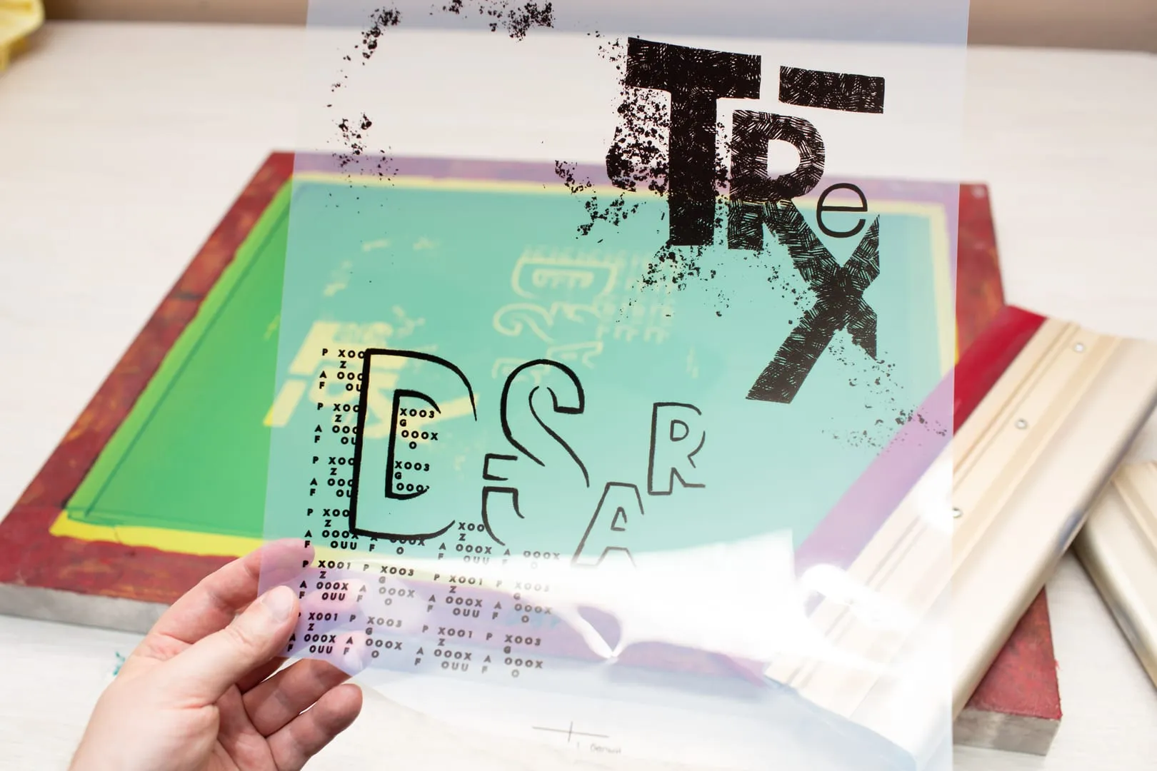 Hand holding a screen-print transparency over a green mesh frame on a studio table