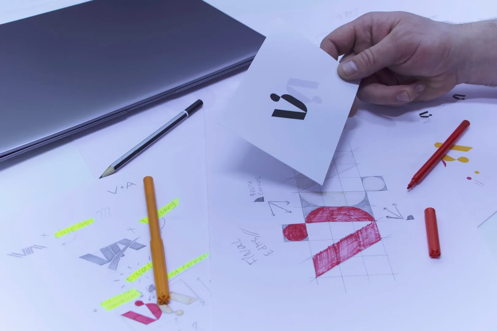 Designer holding a logo mark card over sketch sheets and markers on a studio desk