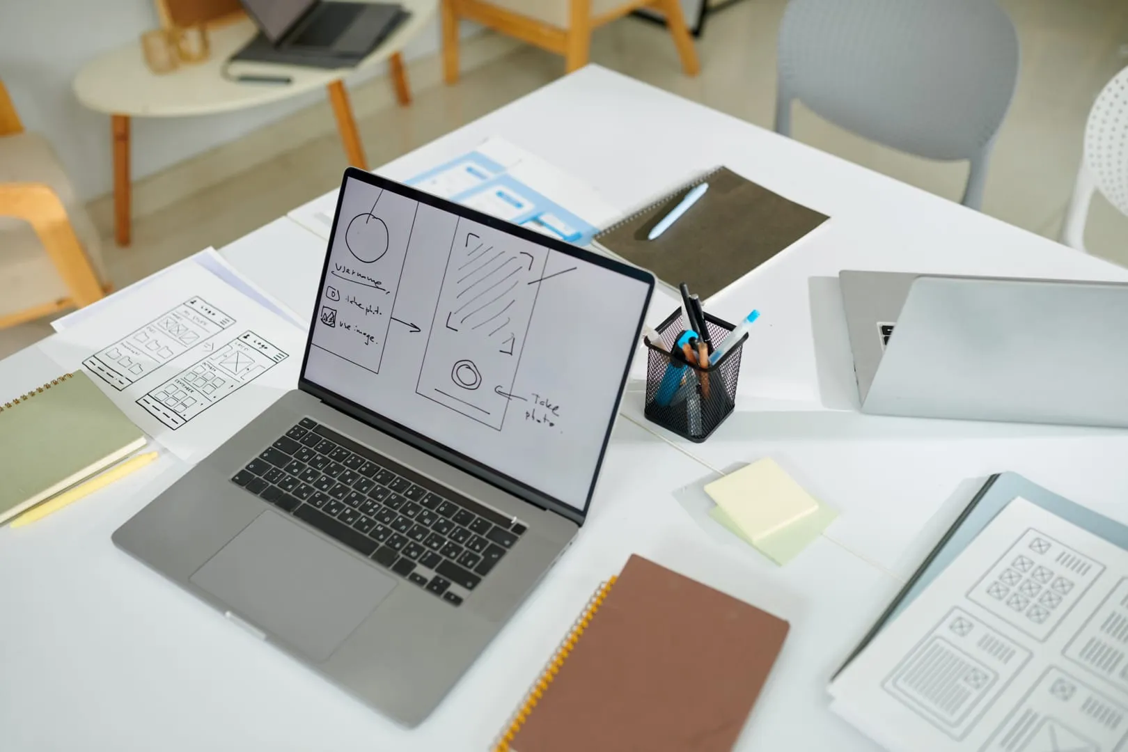 Laptop showing a hand-drawn app wireframe beside paper layouts on a bright studio desk