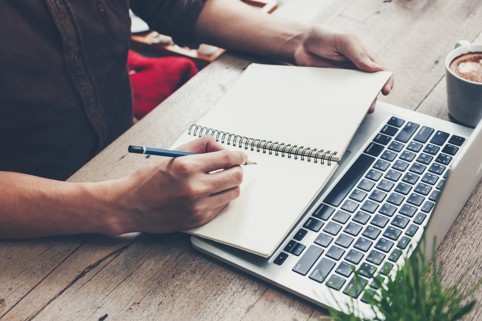 Person writing in a spiral notebook beside a laptop on a wooden desk