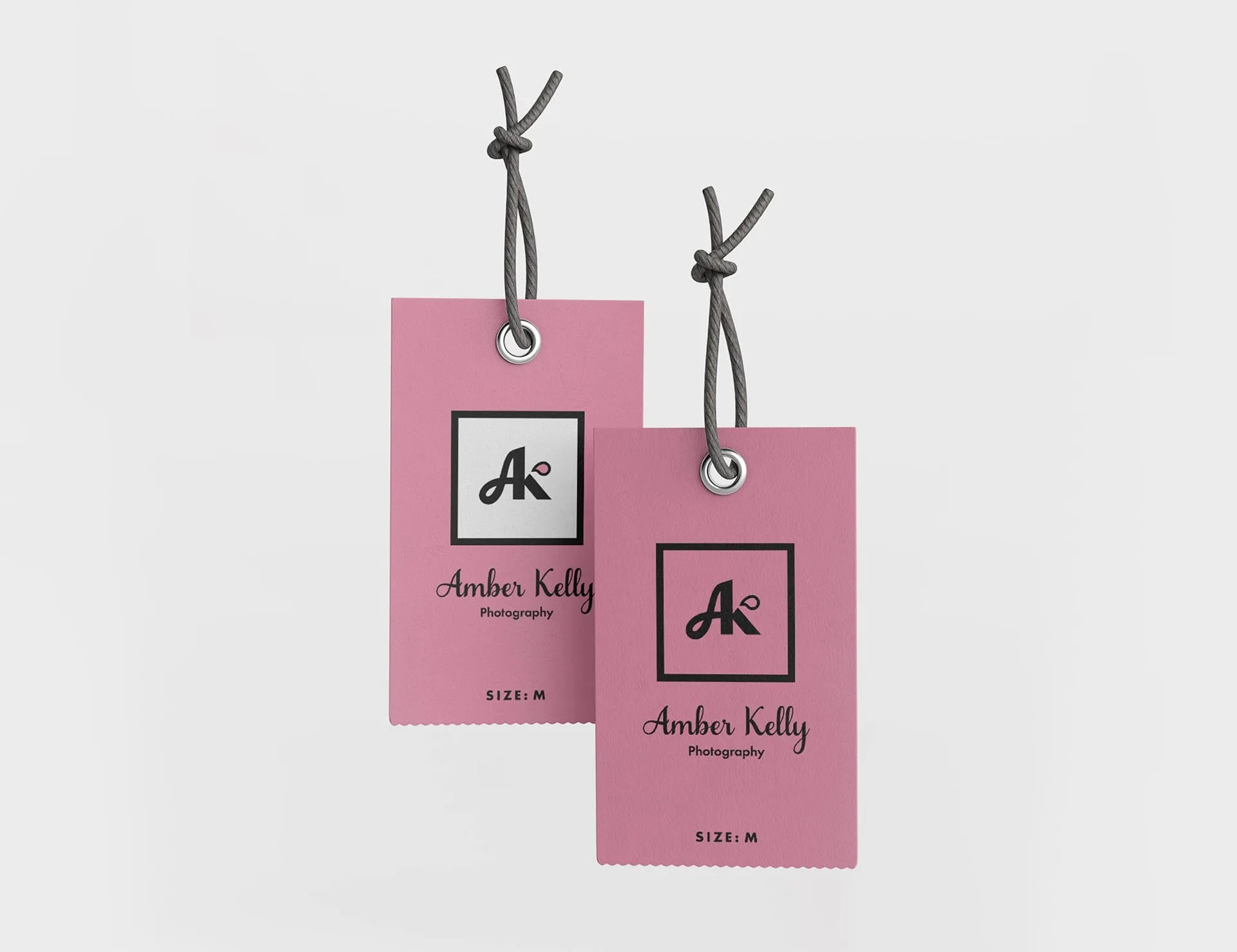 Two pink tags with a logo and text, likely product tags or labels. The tags feature a logo with the letters "AK" in a square, accompanied by the text "Amber Kelly Photography" and "SIZE: M". The tags are attached to a gray string and set ag