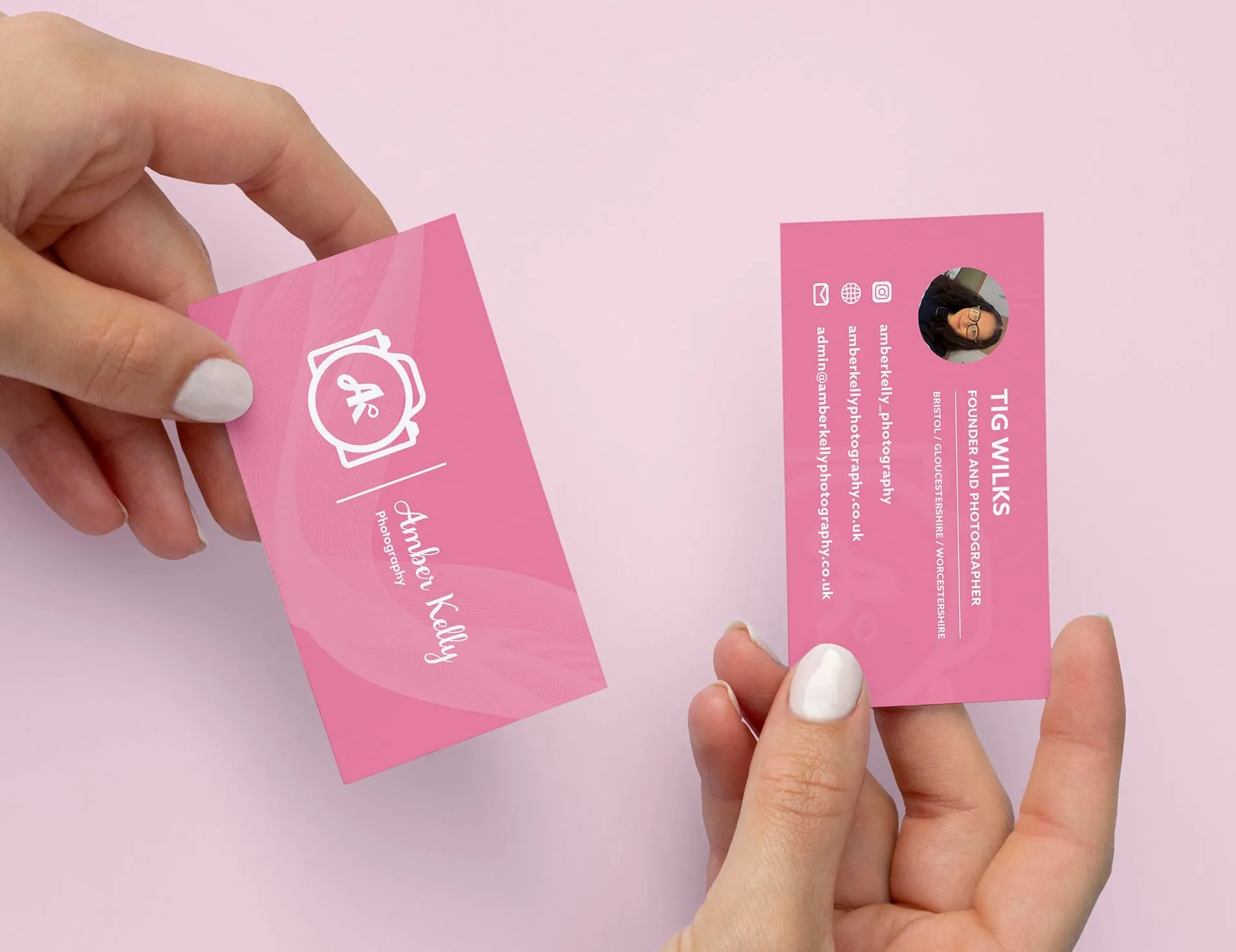 Two hands hold pink business cards, one with a camera logo and the other with a photo of Amber Kelly Photography. The cards are set against a light pink background.