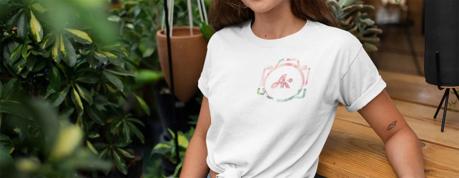 A woman wearing a white t-shirt with a Amber Kelly Photography camera logo, standing in a room with plants and wooden floors.