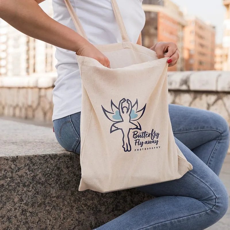 A woman sits on a stone bench, holding a cream-colored tote bag with a logo featuring a stylized butterfly and the words "Butterfly Fly away Photography".