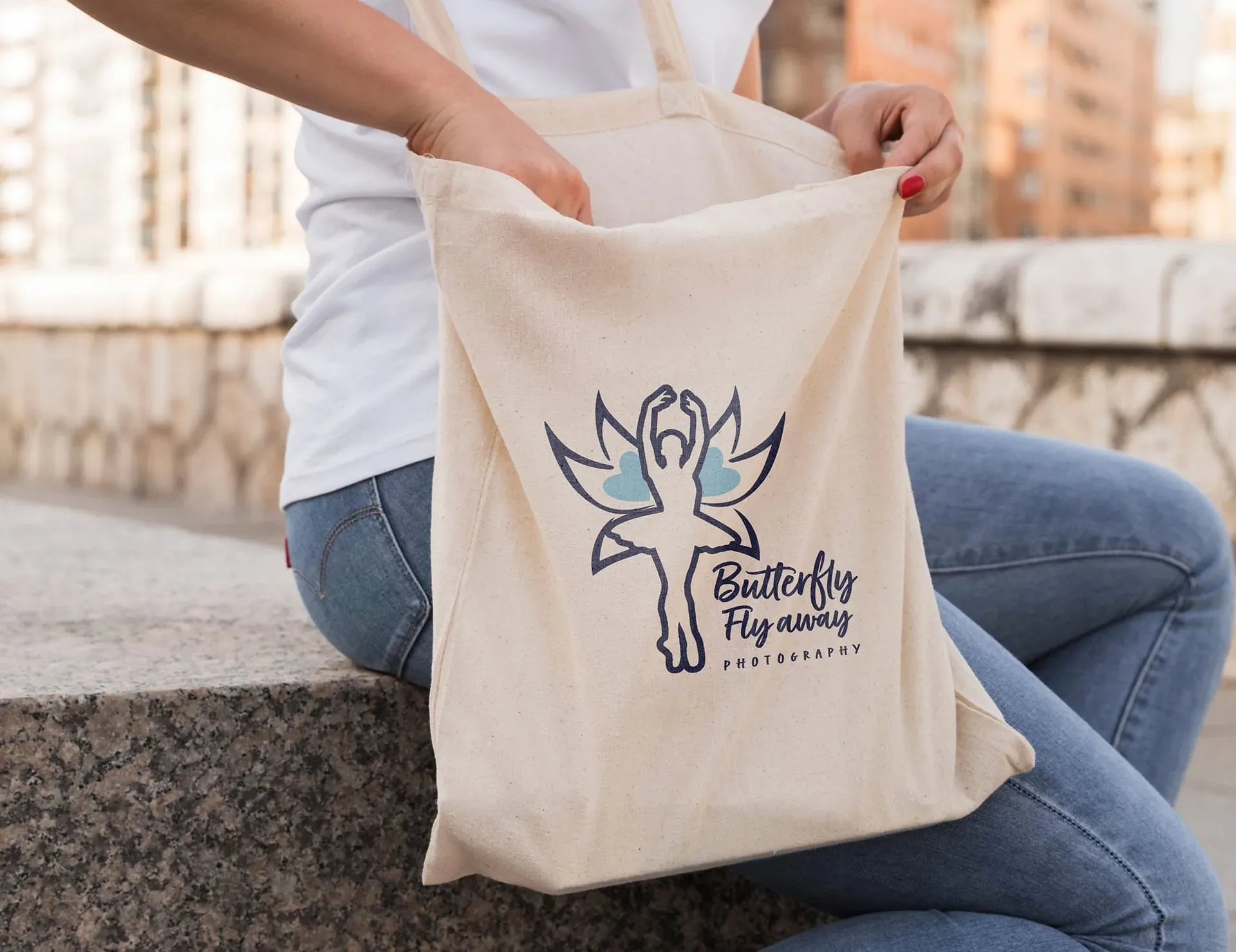 A woman sits on a stone bench, holding a cream-colored tote bag with a logo featuring a stylized butterfly and the words "Butterfly Fly away Photography".