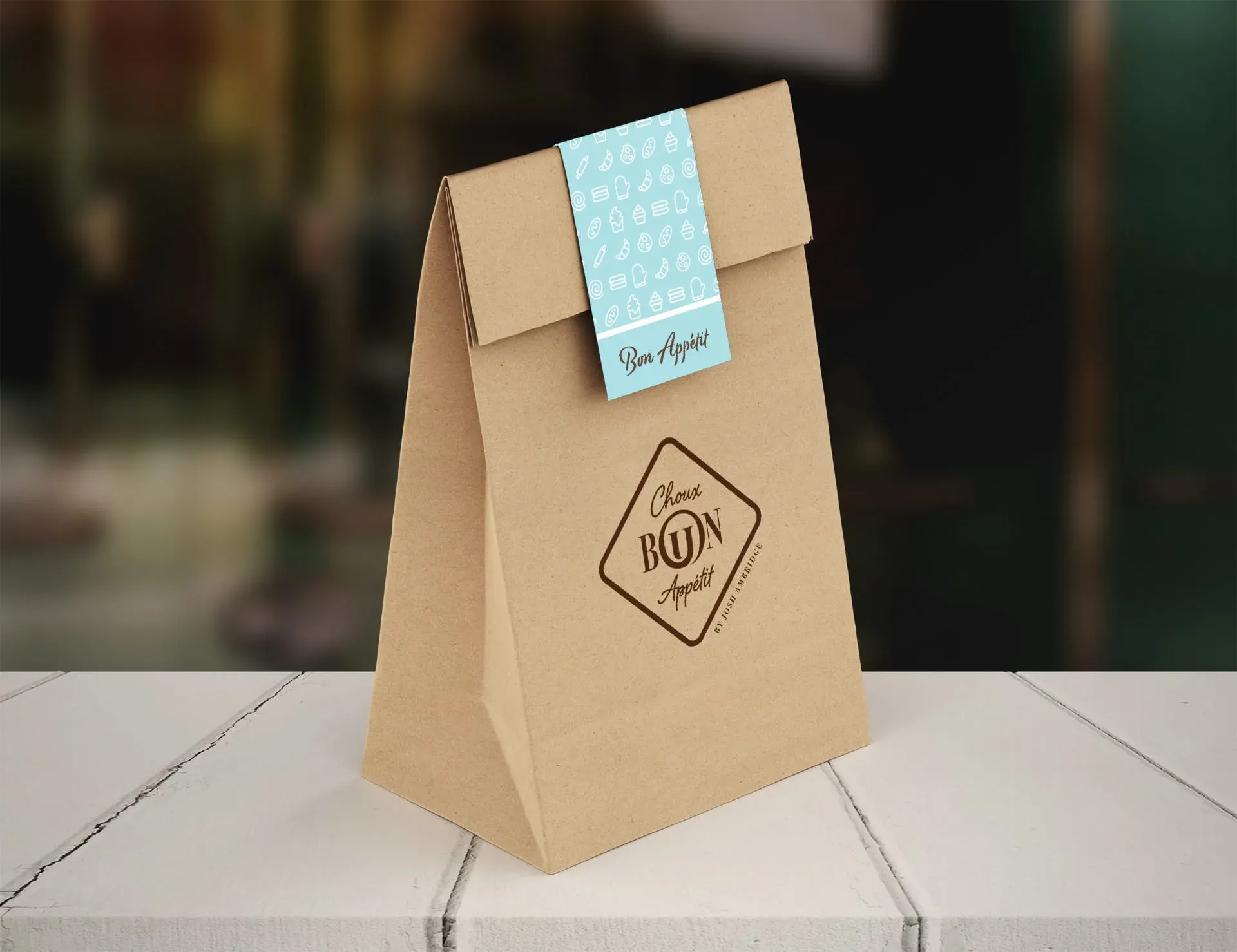 A brown paper bag with a blue label sits on a white wooden surface, likely a table or countertop. The bag features a logo that reads "Choux BUN Appetit" and a blue label with the words "Bon Appetit" in cursive. 
