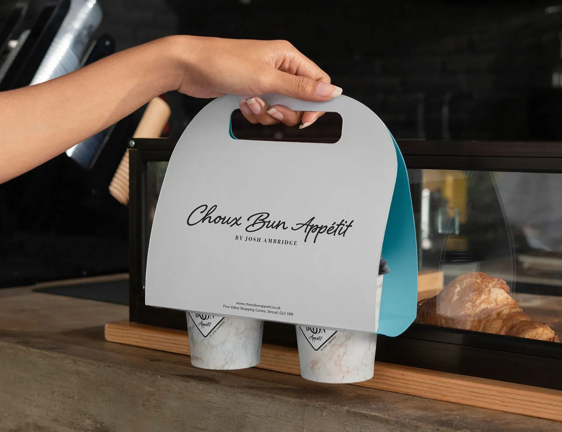A hand holds a white paper bag with a handle, featuring the words "Choux Bun Appetit" in black cursive font. The bag is placed on a wooden counter, with a glass case containing a croissant in the background.