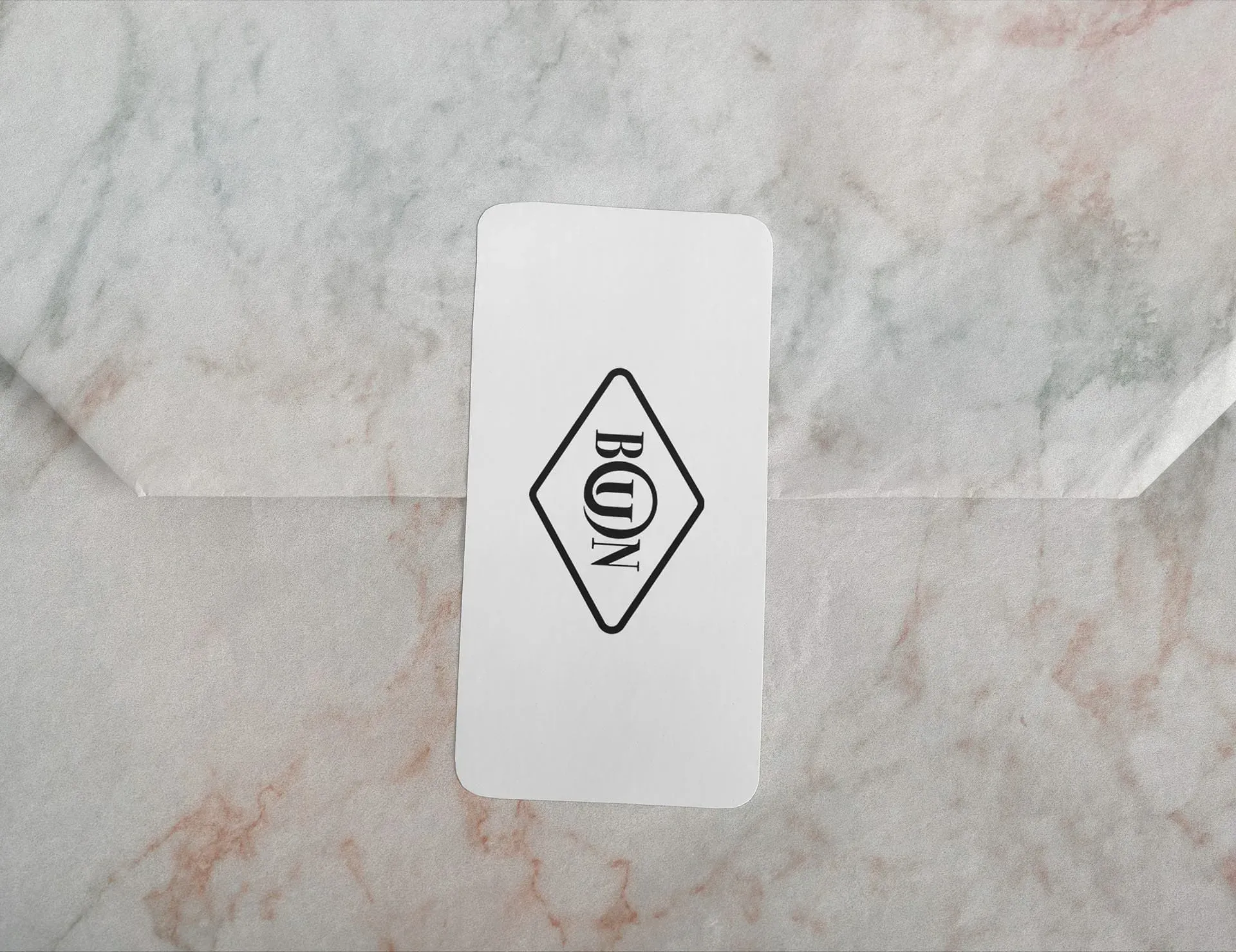 A white business card with a black logo featuring the letters "BON" in a diamond shape, placed on a marble surface. Choux Bun Appétit