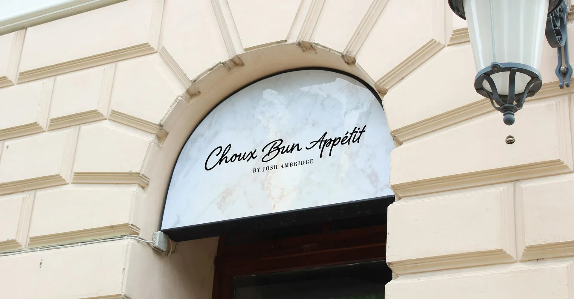 A white marble sign with black text, reading "Choux Bun Appétit by Josh Ambridge", is mounted on a beige wall above an arched doorway. 