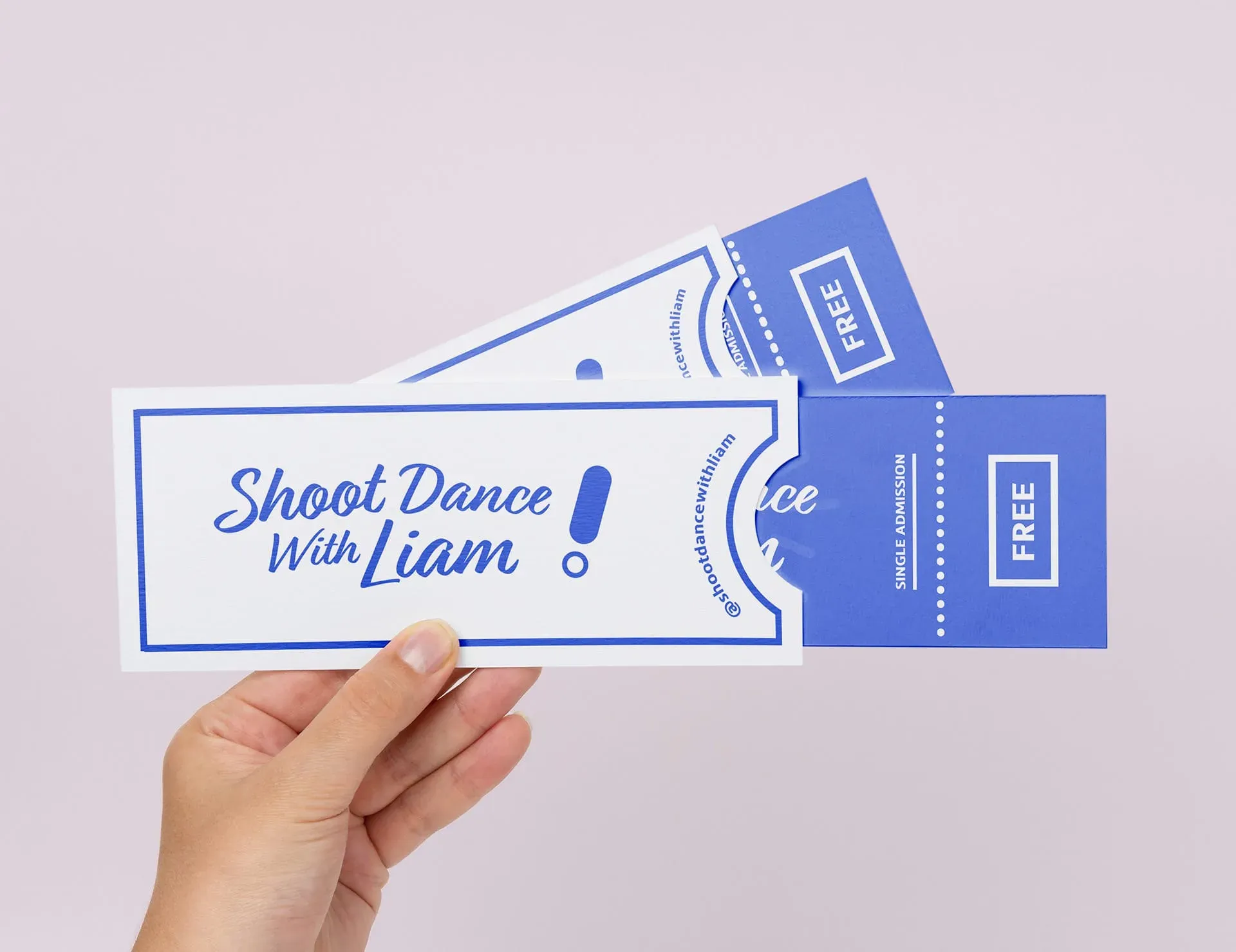 A hand holds a white ticket with blue text reading "Shoot Dance With Liam!" and a blue border. Two blue tickets with white text are visible behind the white ticket. The background is a light pink color.