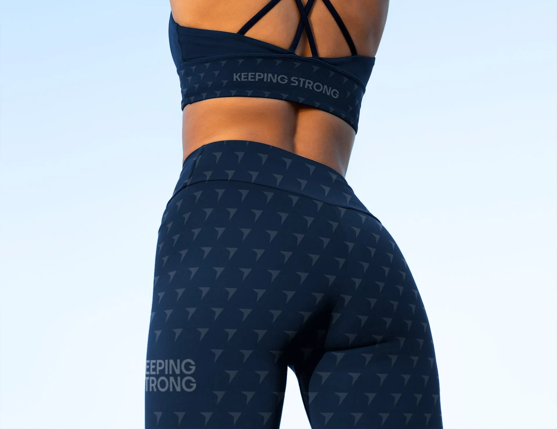 A woman's torso and lower back, wearing a navy blue sports bra and leggings with a repeating pattern of triangles and the words "KEEPING STRONG" printed on the back of the bra and left leg of the leggings.